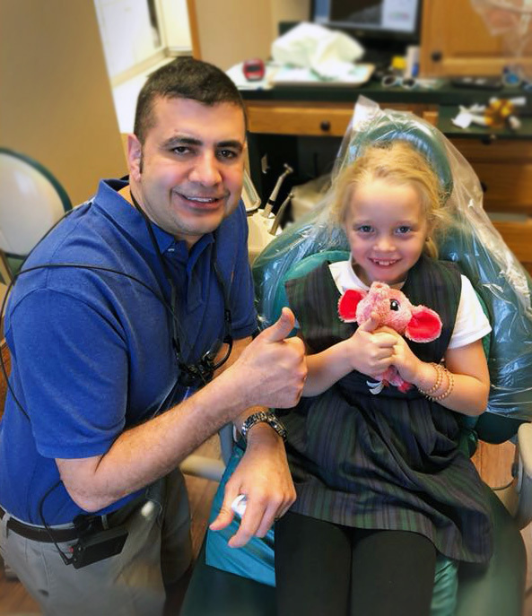 Dr. Neil Bhargava with a young girl in the dentist chair giving a thumbs up